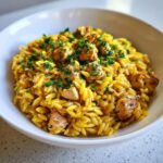 A close-up of a white bowl filled with bright yellow Lemon Chicken Orzo, topped with diced chicken and fresh parsley.