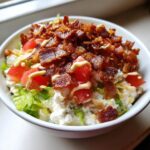 A white bowl filled with a Low Carb BLT Cottage Cheese Bowl, topped with crispy bacon, diced tomatoes, and a drizzle of dressing.