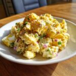 A close-up of creamy Mustard Potato Salad mixed with chunks of egg, red onion, and green onions, sprinkled with paprika.