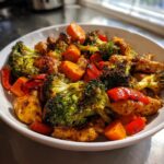 Close-up of roasted One Pan Chicken And Veggies featuring charred broccoli, bright orange carrots, and red peppers in a white bowl.