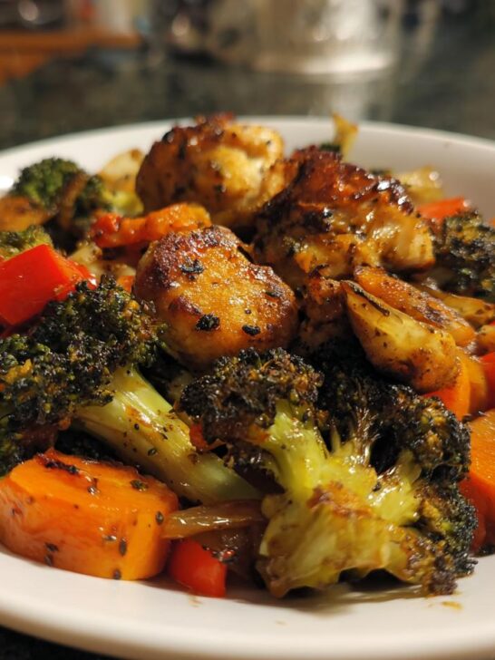 Close-up of a white bowl filled with One Pan Chicken And Veggies, featuring seared chicken, roasted broccoli, carrots, and red peppers.