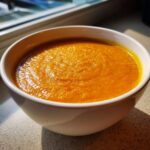 A close-up of a white bowl filled with vibrant orange One Pot Creamy Carrot Lentil Soup, sitting near a window.