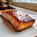 A perfectly cooked fillet of Orange Glazed Salmon resting on a white plate, showing a shiny, caramelized glaze.