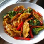Close-up of a serving of Potsticker Stir Fry featuring pan-fried dumplings coated in a glossy sauce with bright green broccoli and red pepper strips.