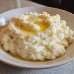 A close-up of fluffy Roasted Garlic Mashed Potatoes in a white bowl, topped with melted butter and black pepper.