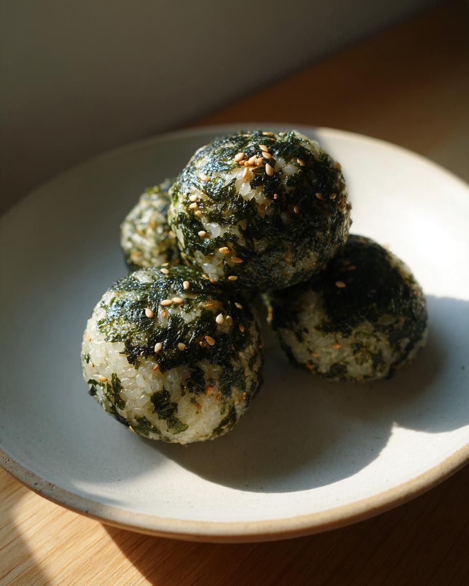 Four perfectly formed Korean Rice Balls coated in seaweed flakes and sprinkled with sesame seeds, resting on a light plate.