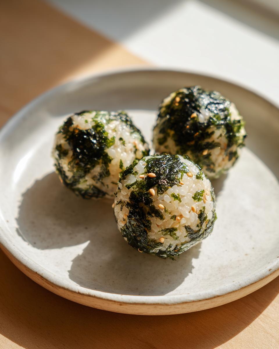 Three perfectly formed Korean Rice Balls coated in seasoned seaweed flakes and sesame seeds on a light ceramic plate.