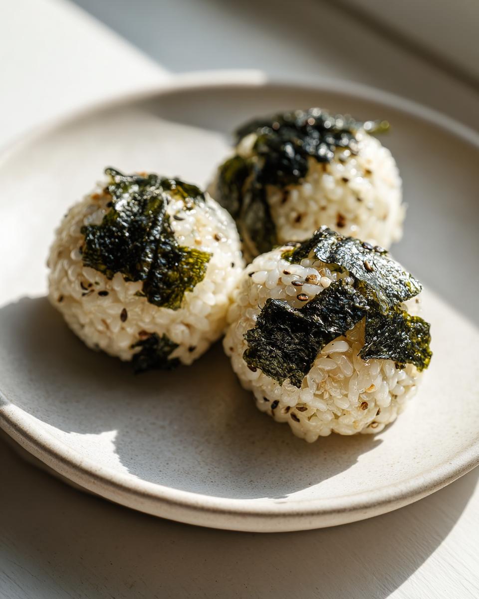 Three perfectly round Korean Rice Balls seasoned with sesame seeds and topped with dark, crispy seaweed, resting on a light plate.