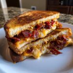 Two halves of a Sun Dried Tomato Bacon Pesto Grilled Cheese stacked, showing melted cheese and bacon.