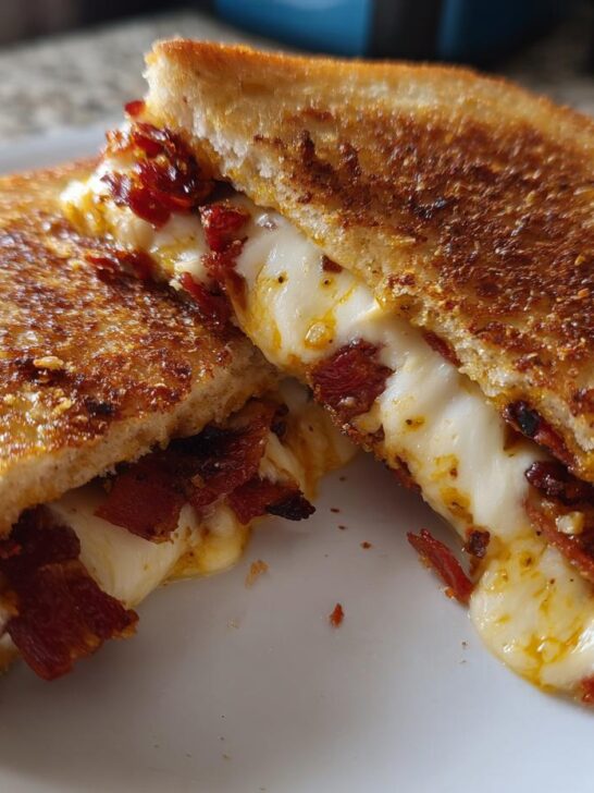 Close-up of a grilled cheese sandwich cut in half, oozing with melted white cheese, bacon, and sun dried tomato bacon pesto.