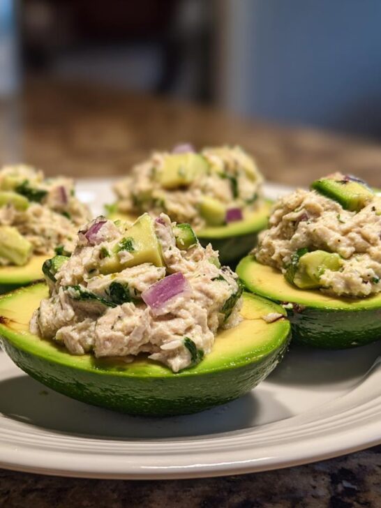 Four halves of avocado filled with creamy tuna salad, known as Tuna Avocado Boats, served on a white plate.